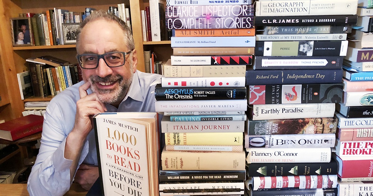 James Mustichevent sitting in a library with stacks of books on the table in front of him